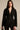 A woman with long brown hair wears the CORTEZ PEPLUM BLAZER in sleek suiting fabric, layered cross necklaces, hoop earrings, and lace-patterned pants, standing against a neutral background and looking at the camera.