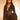 A woman with wavy brown hair wears the CORTEZ PEPLUM BLAZER in black, featuring suiting fabric and a metallic zip. She pairs it with a statement necklace and sheer black stockings, standing confidently against a warmly lit background.