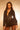 A woman with wavy brown hair wears the CORTEZ PEPLUM BLAZER in black, featuring suiting fabric and a metallic zip. She pairs it with a statement necklace and sheer black stockings, standing confidently against a warmly lit background.