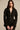 A woman with long brown hair wears the CORTEZ PEPLUM BLAZER, crafted in sleek suiting fabric with a sculpted fit, front zipper, and lace-trimmed sleeves. She layers silver necklaces and stands against a beige background.