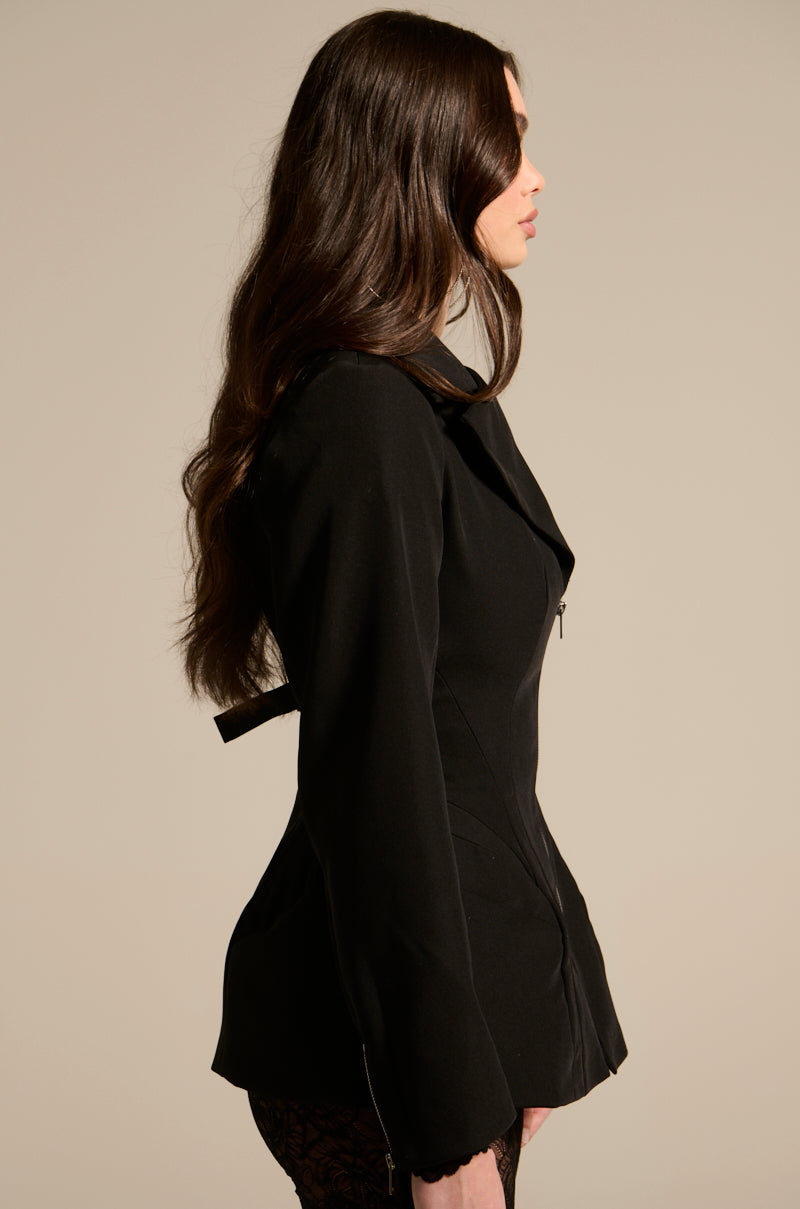 Woman with long brown hair wears the CORTEZ PEPLUM BLAZER in sleek suiting fabric with lace-patterned bottoms, standing in profile against a plain beige background for a sculpted, polished look.