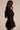 Woman with long brown hair wears the CORTEZ PEPLUM BLAZER in sleek suiting fabric with lace-patterned bottoms, standing in profile against a plain beige background for a sculpted, polished look.