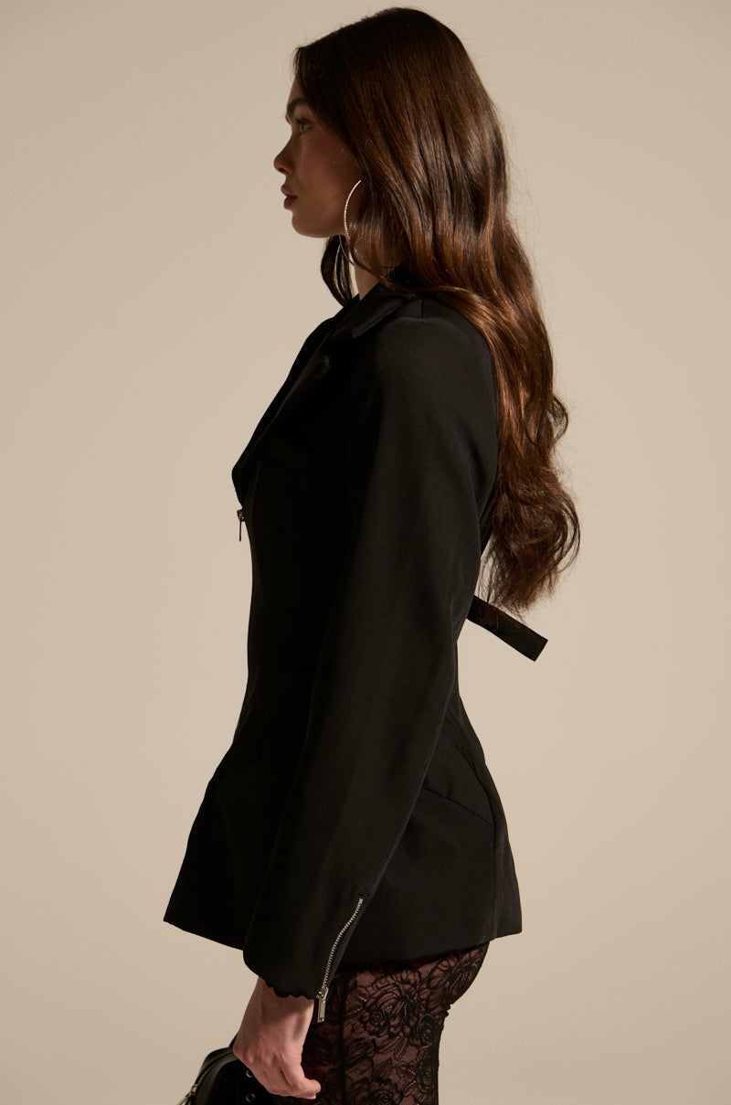 A woman with long wavy brown hair poses in profile against a beige background, wearing the CORTEZ PEPLUM BLAZER, black lace-patterned tights, and holding a small black bag.