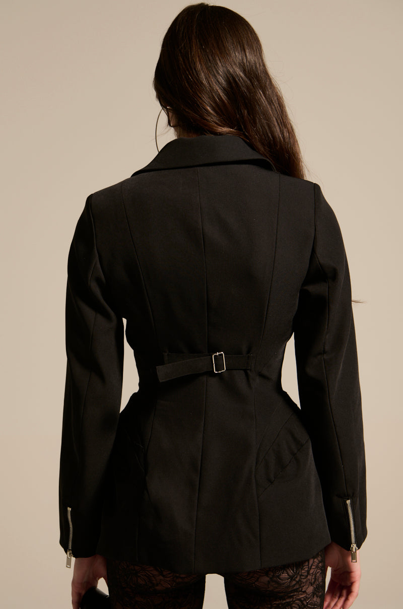 A person with long brown hair is seen from behind, wearing the CORTEZ PEPLUM BLAZER featuring a waist belt, two back vents, and metallic zippered cuffs. The background is plain and neutral.