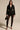 A woman stands facing forward in the CORTEZ PEPLUM BLAZER, styled with black lace leggings, high-heeled sandals, a small black handbag, a silver necklace, and her long hair worn loose.