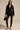 A woman with long hair and a neutral expression stands against a plain background wearing the CORTEZ PEPLUM BLAZER with metallic zippered cuffs, sheer black lace pants, black high-heeled sandals, and holding a small handbag.