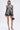 A woman wears the PUSH TO START LACE BLAZER with black shorts, strappy heels, and a small black handbag. She has straight shoulder-length hair, hoop earrings, and stands against a plain background.
