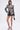 A woman poses against a white background wearing the PUSH TO START LACE BLAZER as a statement piece with a deep neckline, black shorts, strappy black heels, hoop earrings, and a small black handbag.