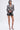 A woman models the PUSH TO START LACE BLAZER with black shorts and strappy high-heeled sandals, holding a small black purse. She stands against a plain white background with sleek, shoulder-length hair.
