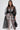 A woman stands confidently against a white background wearing the CAN I KICK IT ORGANZA TRENCH—a sheer black belted dress—over white lace, accessorized with a black handbag, gold earrings, and her long brown hair.