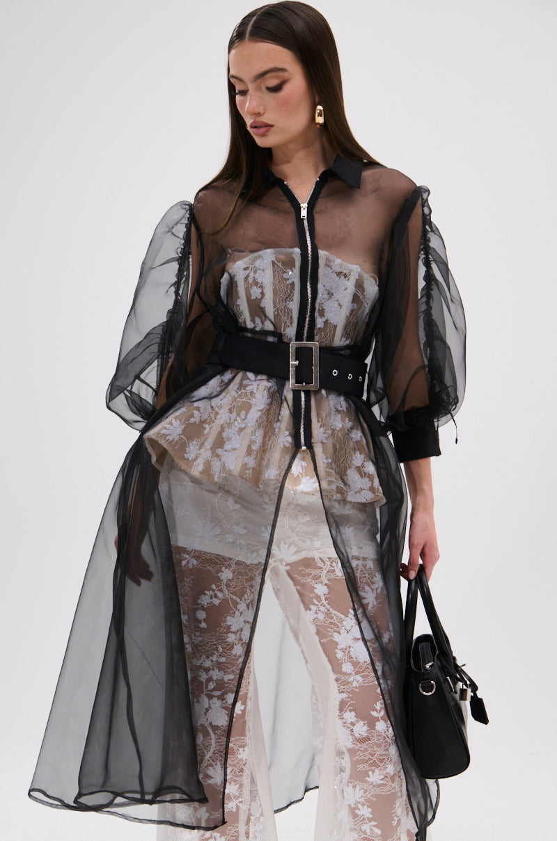 A woman wears the CAN I KICK IT ORGANZA TRENCH with puff sleeves and a wide belt over a white lace top and pants, holding a black handbag. The A-line silhouette pops against the plain, light-colored background as she looks downward.