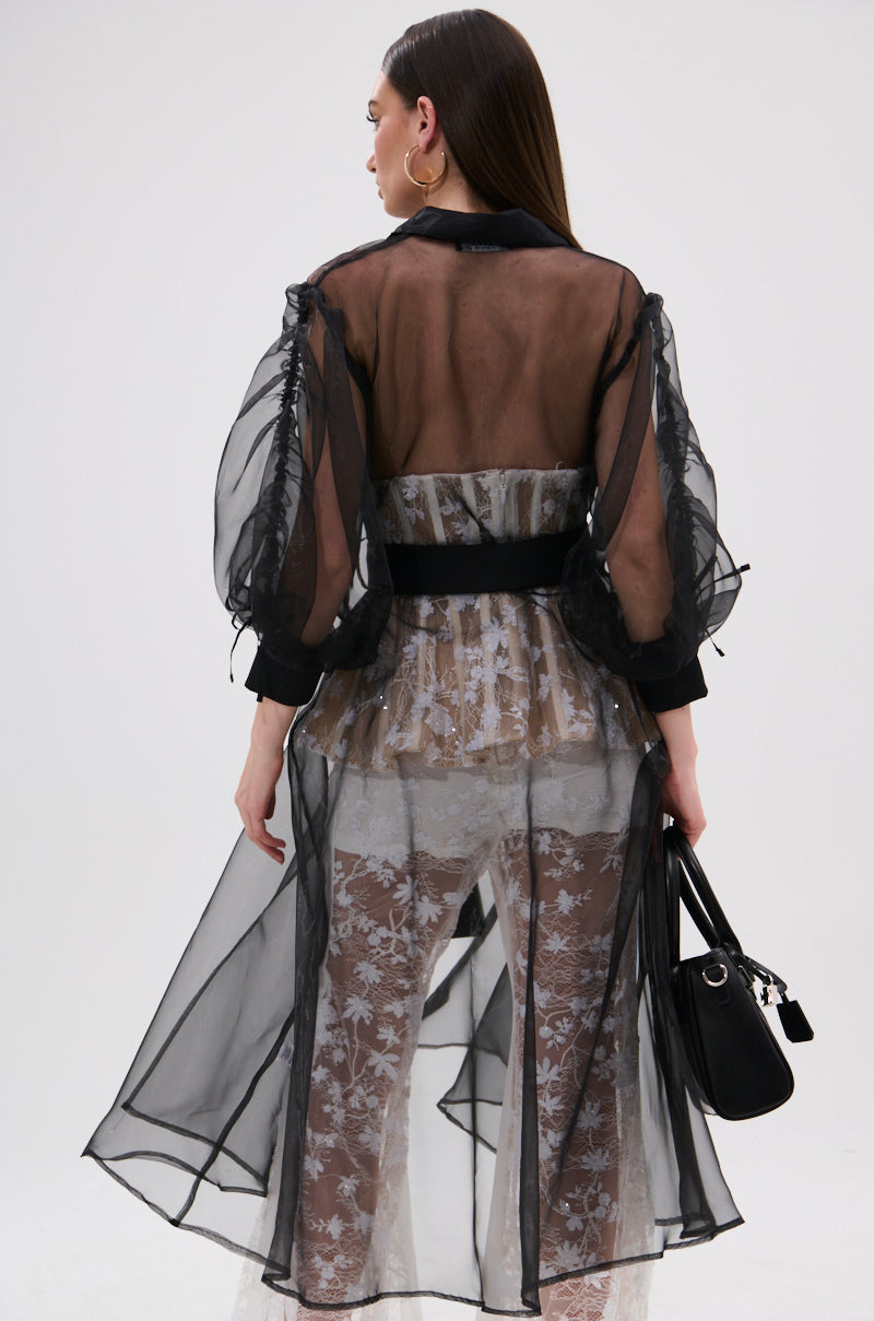 A woman with straight brown hair stands facing away in the CAN I KICK IT ORGANZA TRENCH, showcasing its voluminous sleeves over a beige, white floral lace outfit. She holds a black handbag against a plain light background.
