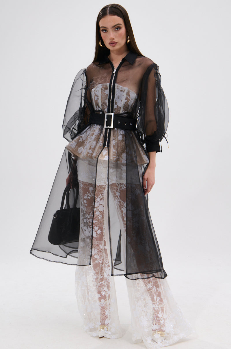 A woman stands against a white background wearing the CAN I KICK IT ORGANZA TRENCH belted over a white lace outfit. She holds a black handbag, has straight brown hair, and wears statement earrings.