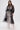 A woman stands against a white background wearing the CAN I KICK IT ORGANZA TRENCH belted over a white lace outfit. She holds a black handbag, has straight brown hair, and wears statement earrings.