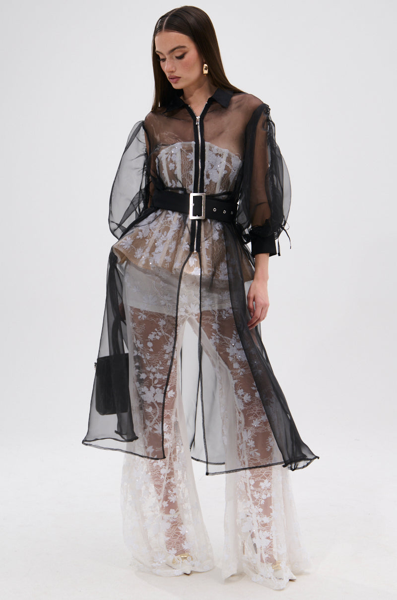 A woman wears the CAN I KICK IT ORGANZA TRENCH—sheer black with puffed long sleeves and a belted waist—layered over a white lace wide-leg jumpsuit, standing against a plain white background.