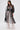 A woman wears the CAN I KICK IT ORGANZA TRENCH—sheer black with puffed long sleeves and a belted waist—layered over a white lace wide-leg jumpsuit, standing against a plain white background.