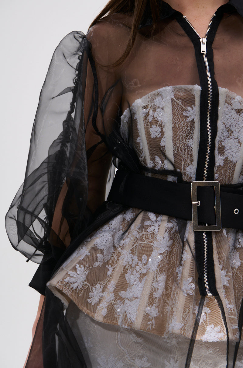 A close-up of a person wearing the CAN I KICK IT ORGANZA TRENCH—a sheer black organza top with puffy sleeves—over a white lace corset and wide black belt with silver buckle, showcasing an A-line silhouette with a front zipper.