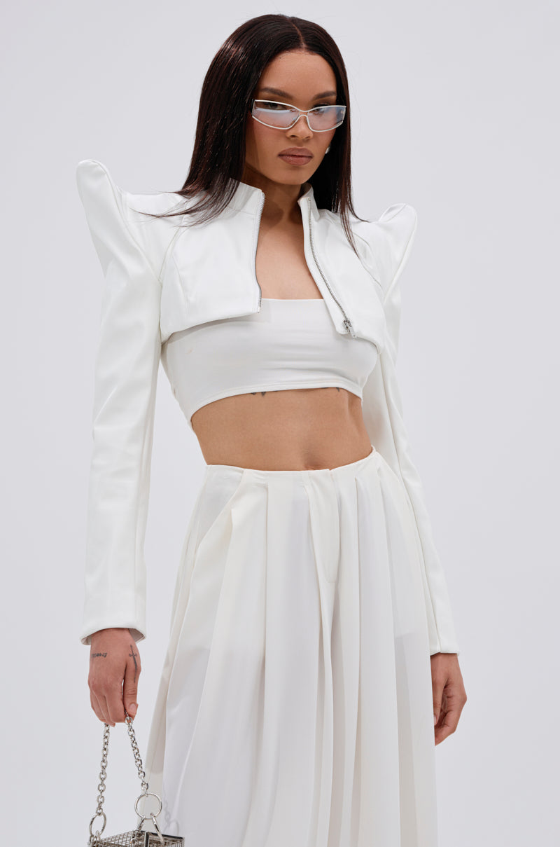 She wears the TAKING NAMES MOTO BOLERO, a white cropped jacket with structured shoulders, over a white bandeau top and flowing skirt. Her look is finished with straight dark hair, clear-framed glasses, and a small chain handbag. 0