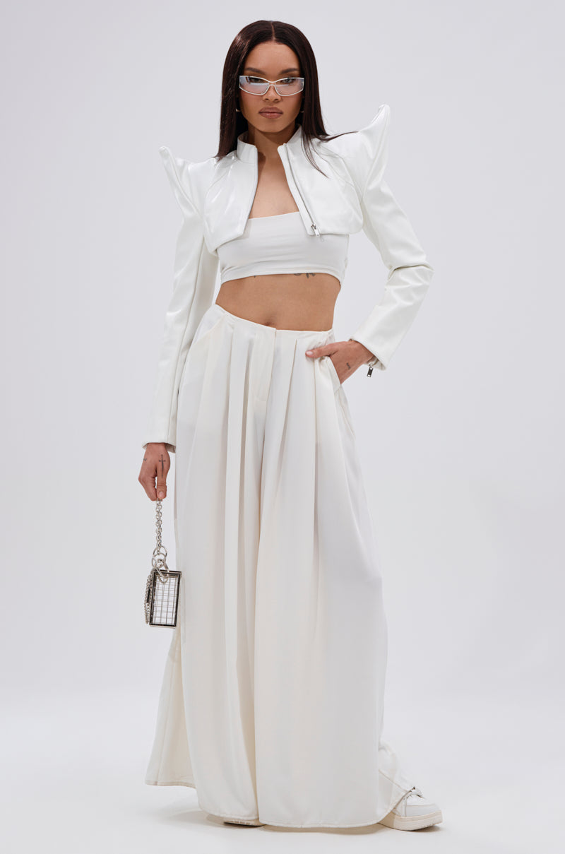 A woman wears the TAKING NAMES MOTO BOLERO with pointed shoulders, paired with futuristic white sunglasses, a white bandeau top, high-waisted wide-leg pants, and holds a small metallic handbag against a plain background. 11