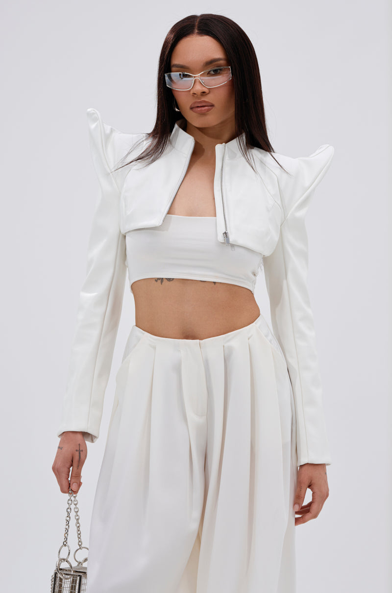 A woman wears the TAKING NAMES MOTO BOLERO—a stylish white cropped jacket with pointed shoulders—and pairs it with a bandeau top, high-waisted pleated pants, a metallic purse, and narrow rectangular sunglasses. 1