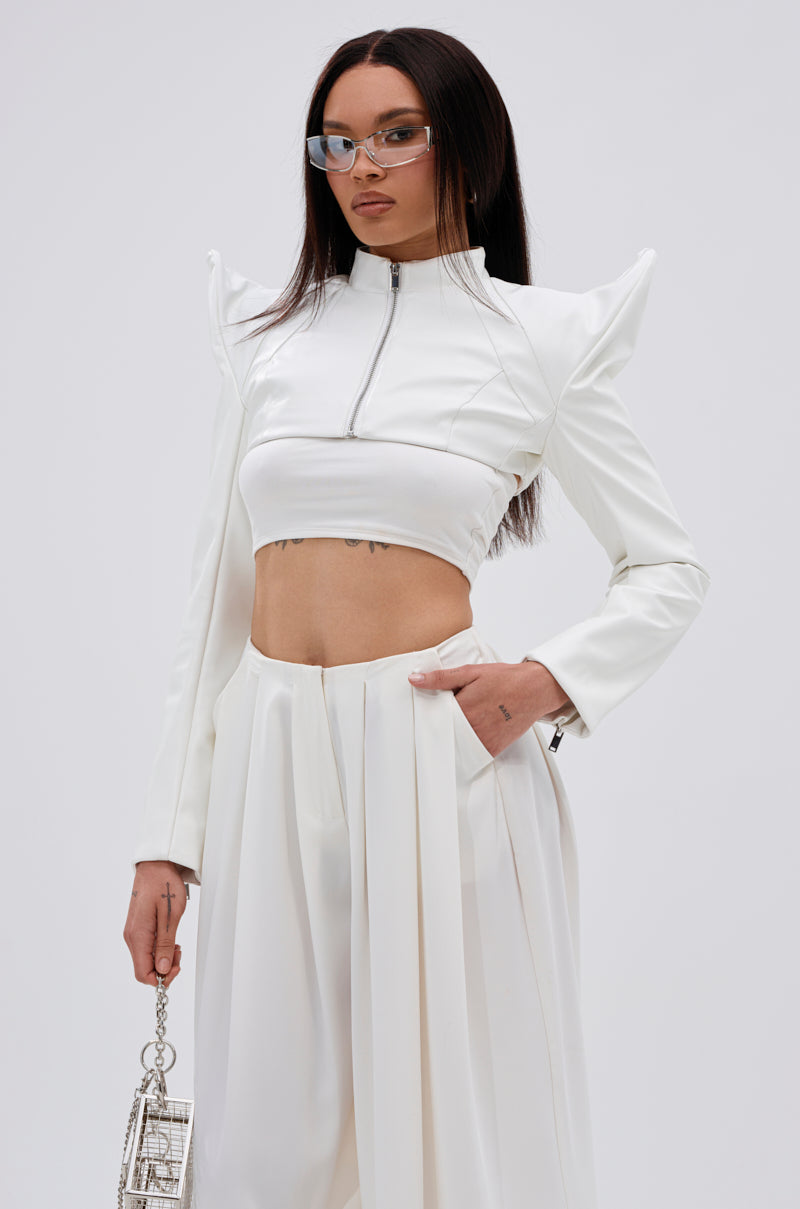 A woman wears the TAKING NAMES MOTO BOLERO, a white cropped moto jacket with structured shoulders, and wide-leg white pants. She has straight hair, glasses, and carries a small silver handbag. 2