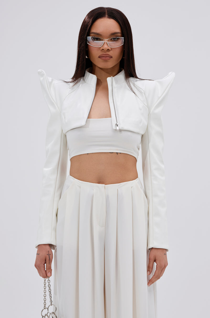 A woman stands against a plain light background in the TAKING NAMES MOTO BOLERO, styled with pointed shoulders, a matching white bandeau top, high-waisted wide-leg pants, and clear rectangular glasses. 3