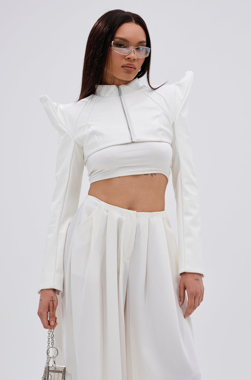 Wearing the TAKING NAMES MOTO BOLERO—a futuristic white cropped jacket with pointed shoulders—and high-waisted pleated white pants, a person accessorizes with a small silver handbag and glasses against a plain background. 4