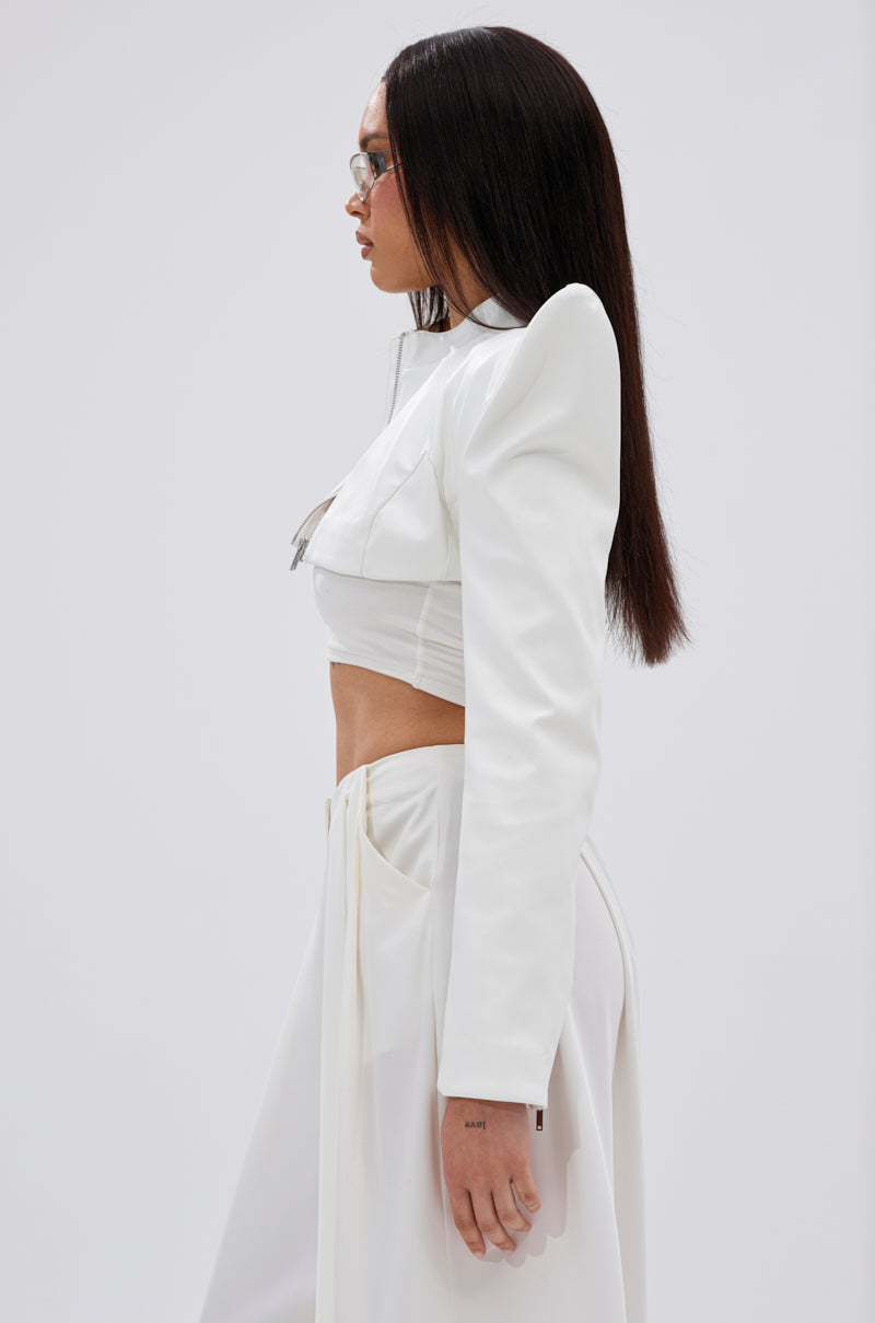 A woman with straight dark hair, glasses, and a stylish white TAKING NAMES MOTO BOLERO with high-waisted pants stands in profile against a plain light background. 5