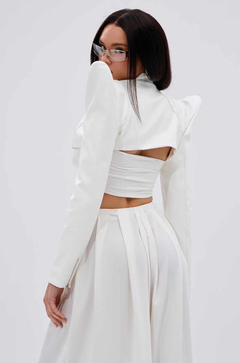 A woman with straight dark hair confidently poses in the TAKING NAMES MOTO BOLERO, a stylish white bolero with structured shoulders and an open back, paired with a flowing skirt against a plain light background. 6