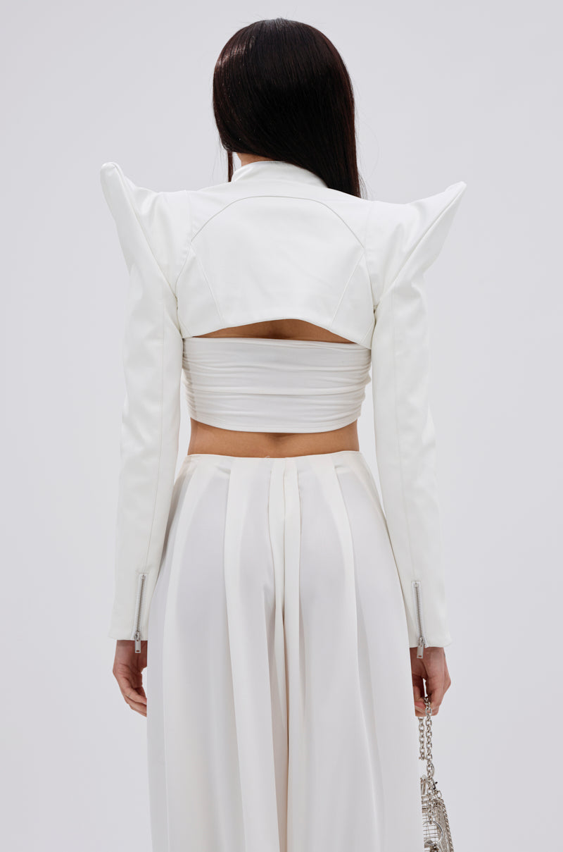 A woman with long dark hair faces away, wearing the TAKING NAMES MOTO BOLERO—a white cropped jacket with pointed shoulders and a cutout back—paired with high-waisted, wide-leg white pants. She holds a small silver handbag in her left hand. 7