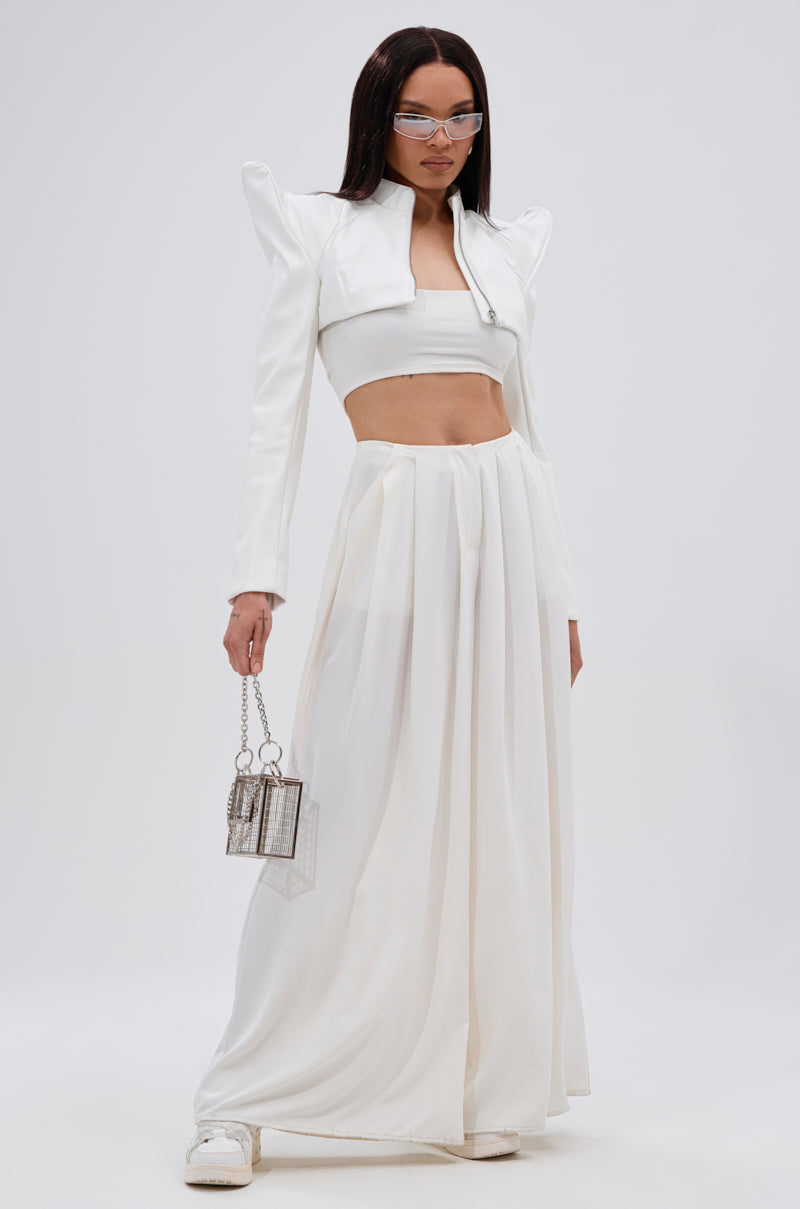 A woman stands against a plain background wearing the TAKING NAMES MOTO BOLERO, a white crop top, long flowing white skirt, white sneakers, clear sunglasses, and holding a small transparent handbag. 8