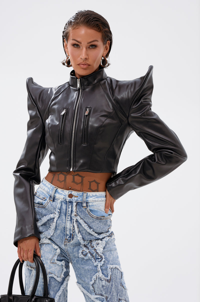 A woman wears the STRAIGHT TO THE POINT MOTO, a black cropped jacket with bold shoulders, paired with high-waisted light-wash textured jeans. She holds a black handbag and poses confidently against a plain background.