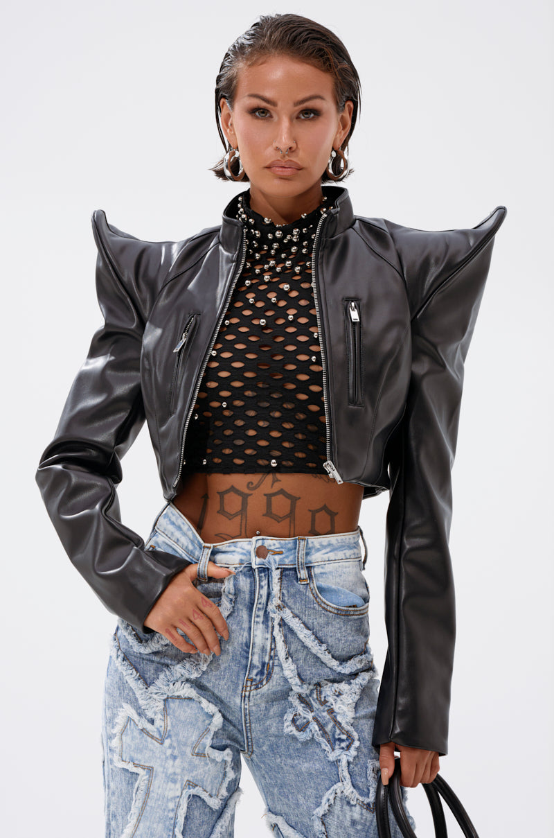Wearing the STRAIGHT TO THE POINT MOTO black cropped faux leather jacket with bold shoulders, a black fishnet crop top, light blue distressed jeans, silver hoop earrings, and holding a black handbag, she stands confidently.