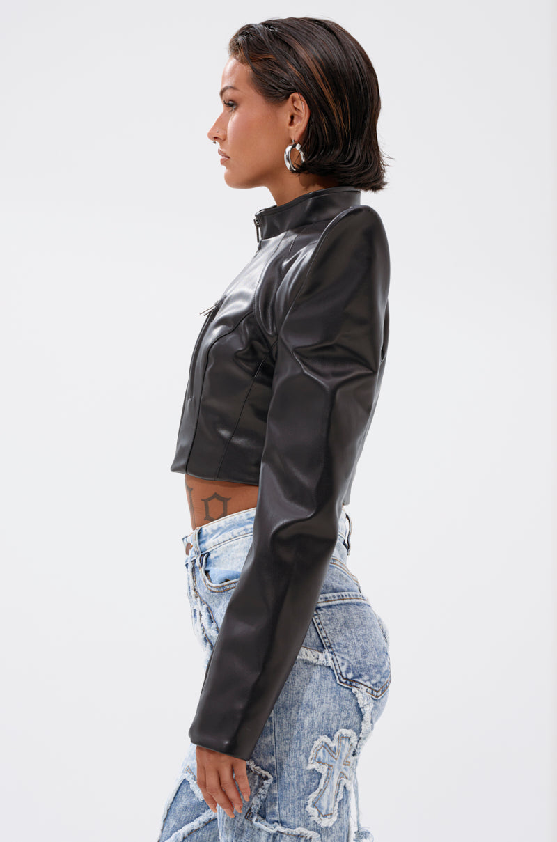 A person with short dark hair and hoop earrings wears the STRAIGHT TO THE POINT MOTO— a cropped black faux leather jacket—paired with light blue distressed jeans featuring cross designs, standing in profile against a plain white background.