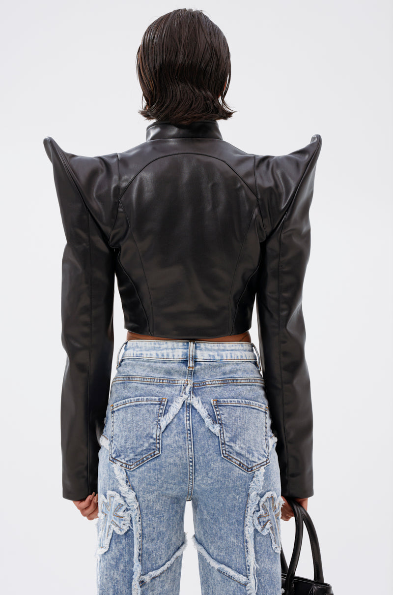 A person with short dark hair is shown from the back, wearing the STRAIGHT TO THE POINT MOTO jacket—black faux leather with pointed shoulders and a cropped fit—paired with light-wash, high-waisted jeans featuring frayed seams and star patches.