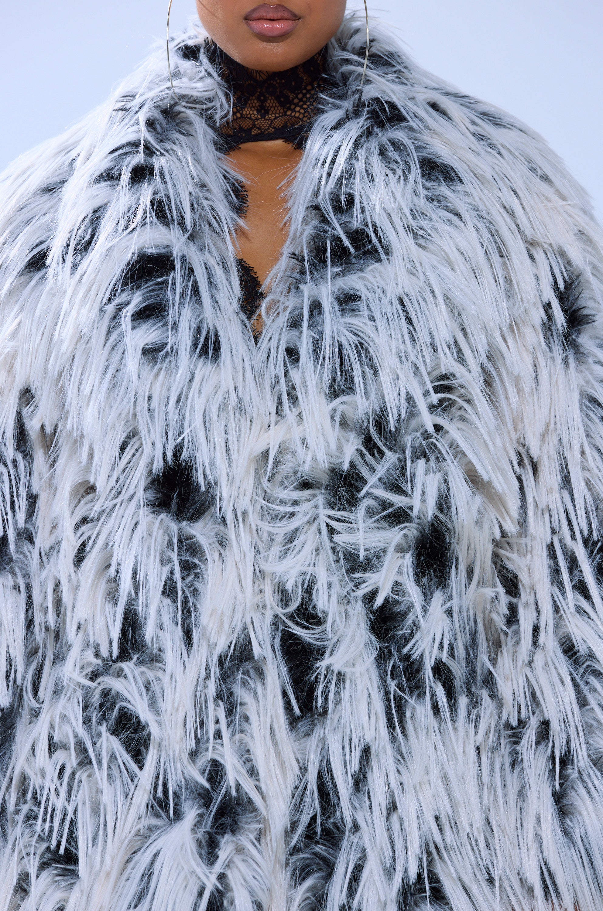 A person wears the EXPENSIVE TASTE FAUX FUR, a thick, longline black and white coat with shaggy fibers; only their lower face, neck, and chest are visible.