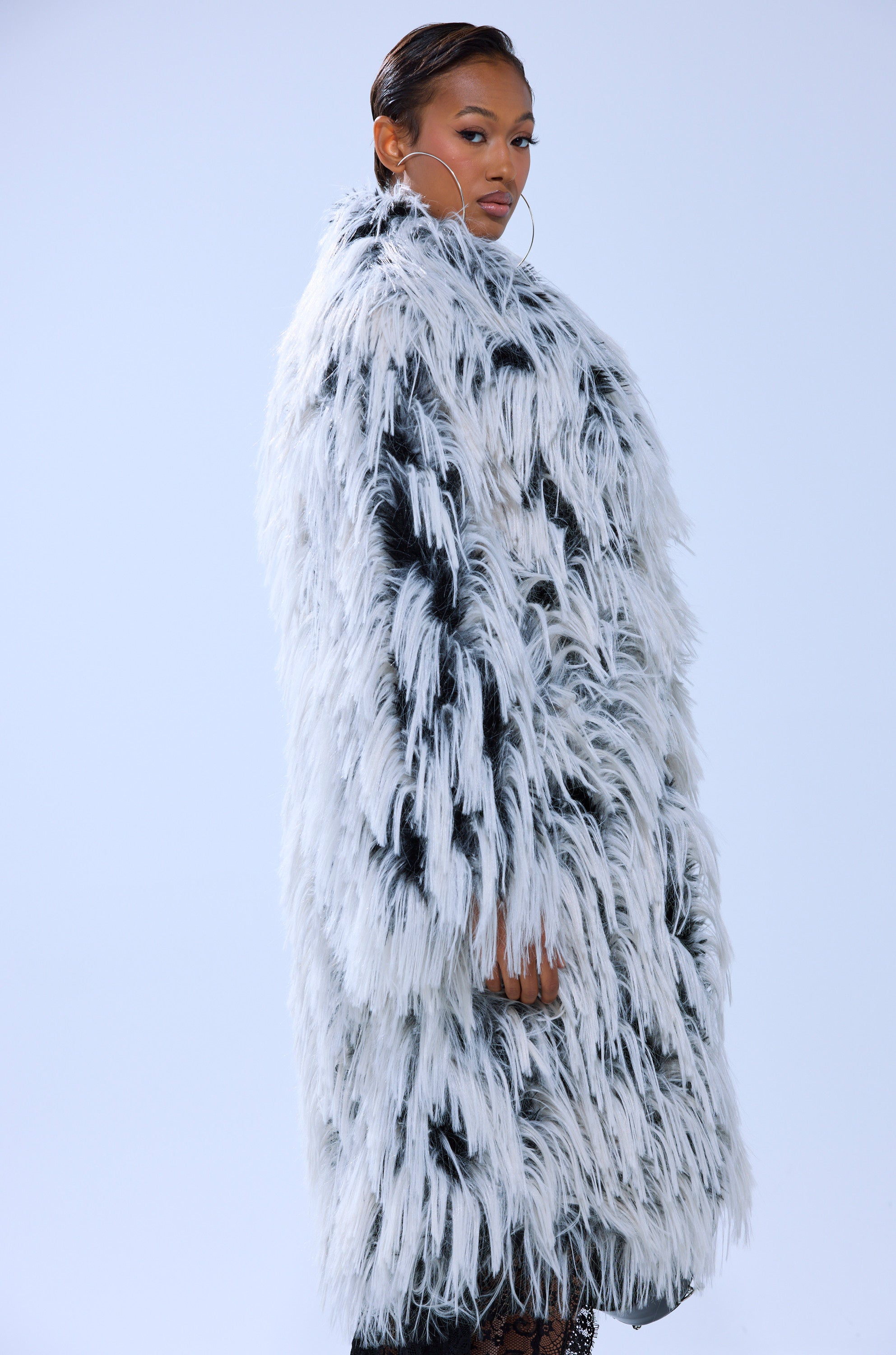 A woman stands sideways, looking at the camera, wearing the EXPENSIVE TASTE FAUX FUR jacket and large hoop earrings against a plain light background.