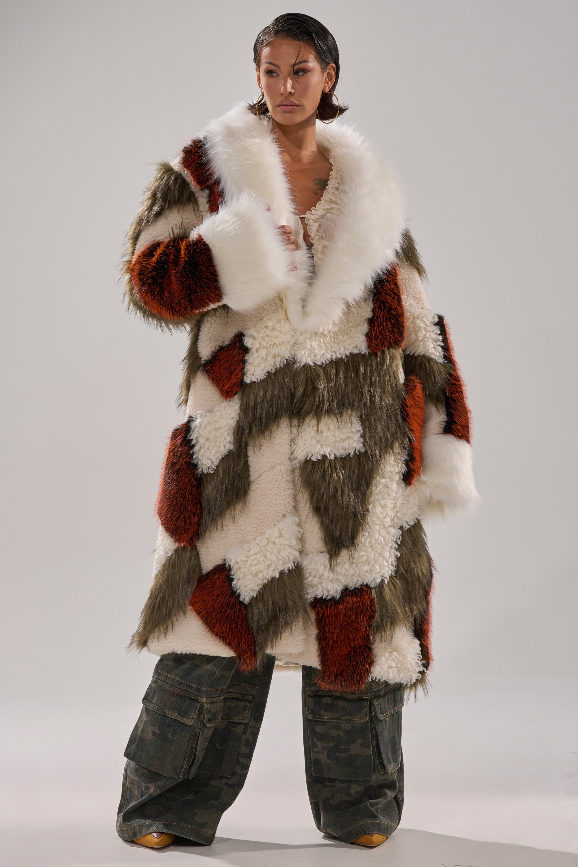 A person stands confidently in the FLEUR PATCHWORK FAUX FUR coat—cream, brown, and rust—paired with baggy camo pants and tan shoes against a plain background.
