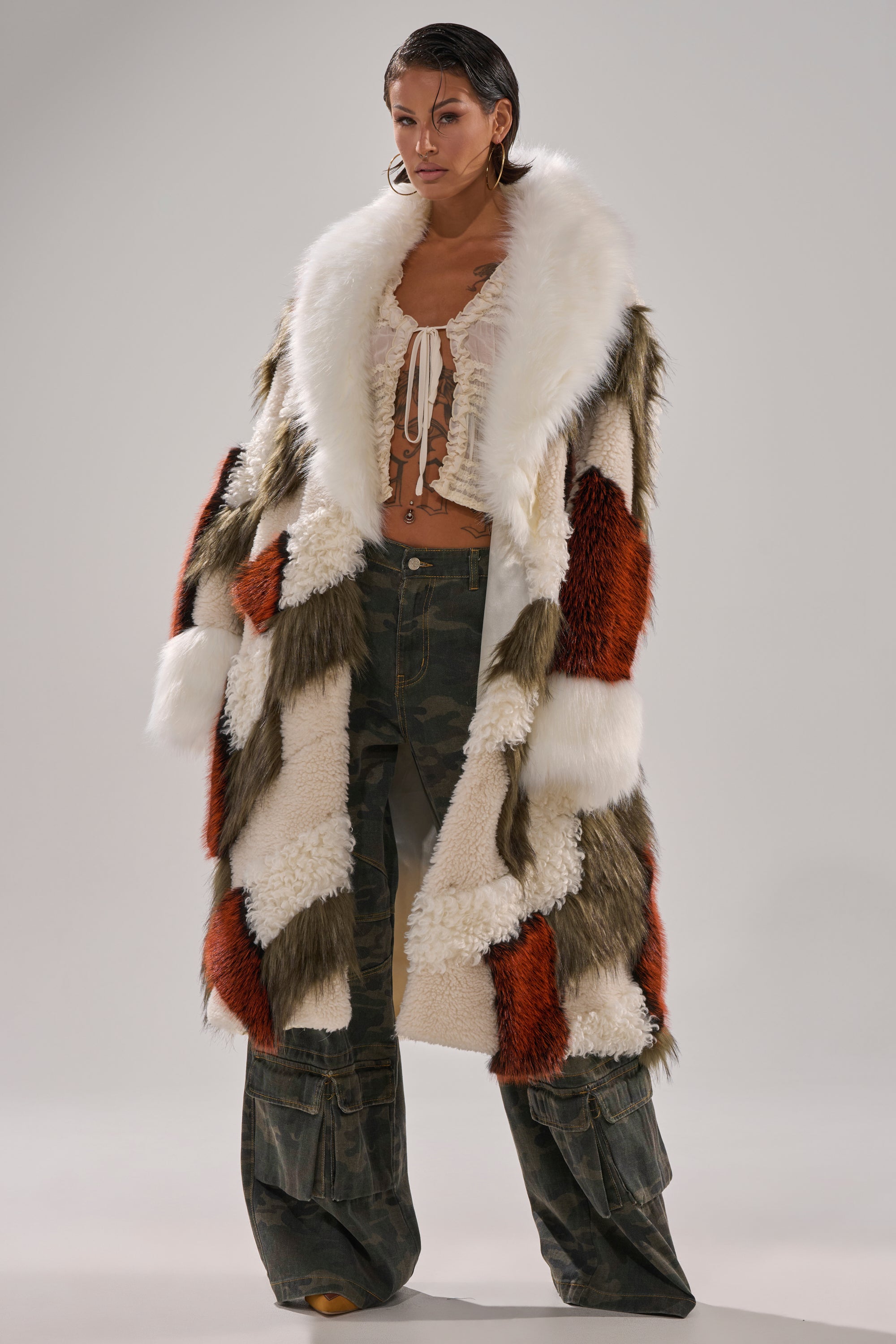 A person poses in the FLEUR PATCHWORK FAUX FUR coat with white, olive, and rust panels over a ruffled white crop top and wide-leg camo pants, set against a plain light background.
