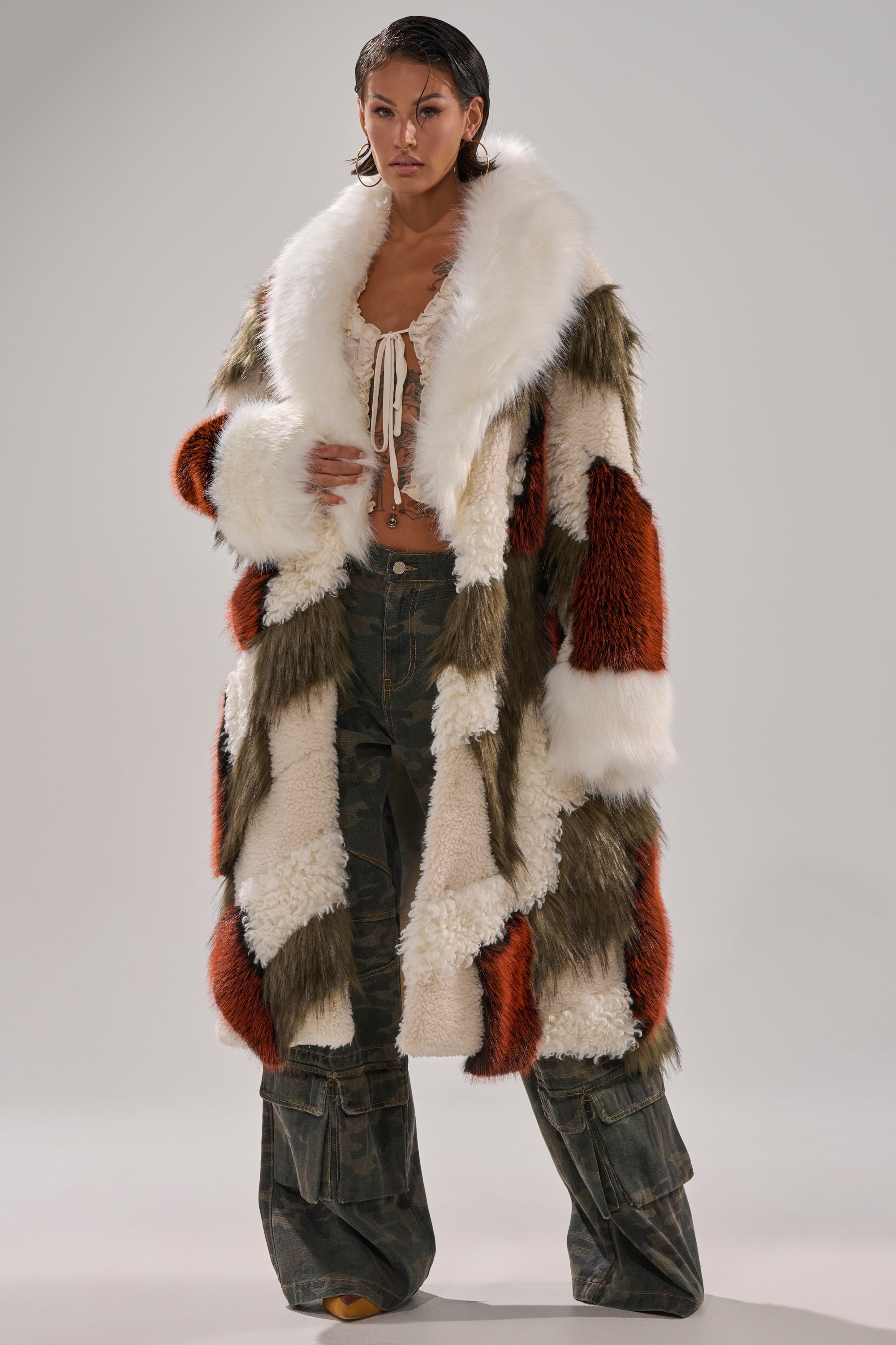 Wearing the FLEUR PATCHWORK FAUX FUR coat in white, orange, and green tones with a cropped cream top and loose camouflage pants, a person stands against a plain, light background.
