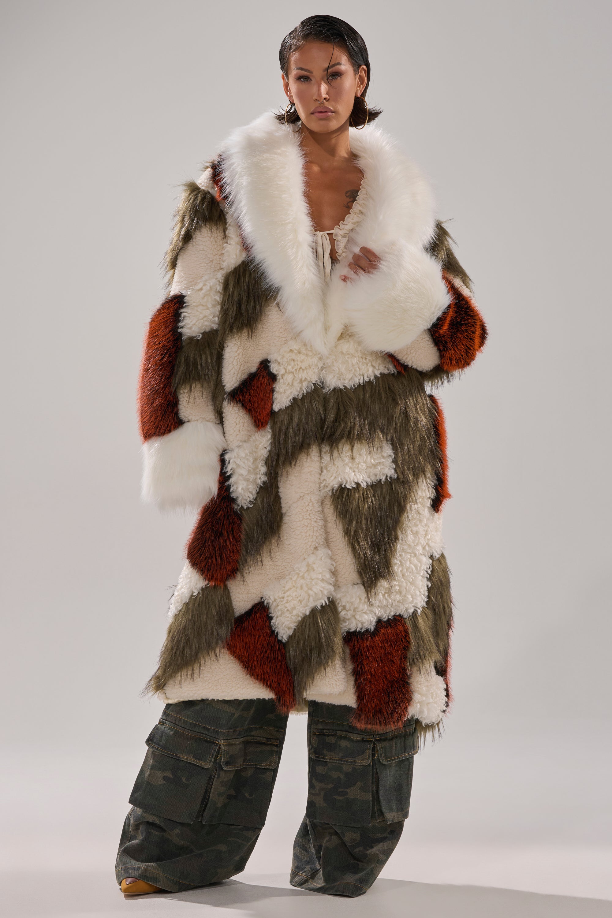 A person stands confidently in the FLEUR PATCHWORK FAUX FUR coat with white, olive, and rust panels and a large white collar. They have slicked-back hair, loose camouflage cargo pants, and pose against a plain light background.