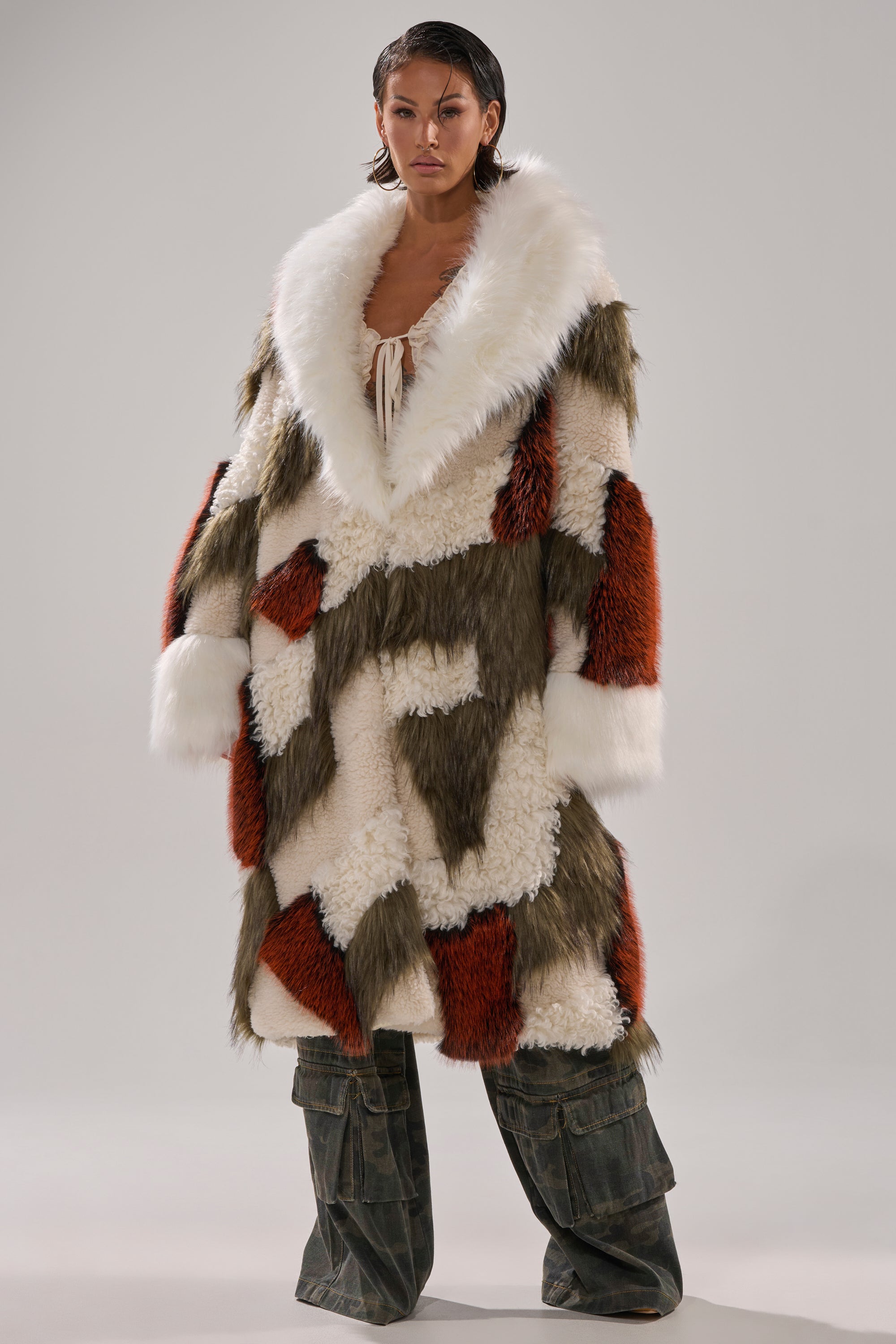 A person stands against a plain background wearing the FLEUR PATCHWORK FAUX FUR coat featuring white, brown, and olive green panels, wide sleeves, a large white collar, and paired with loose dark pants.