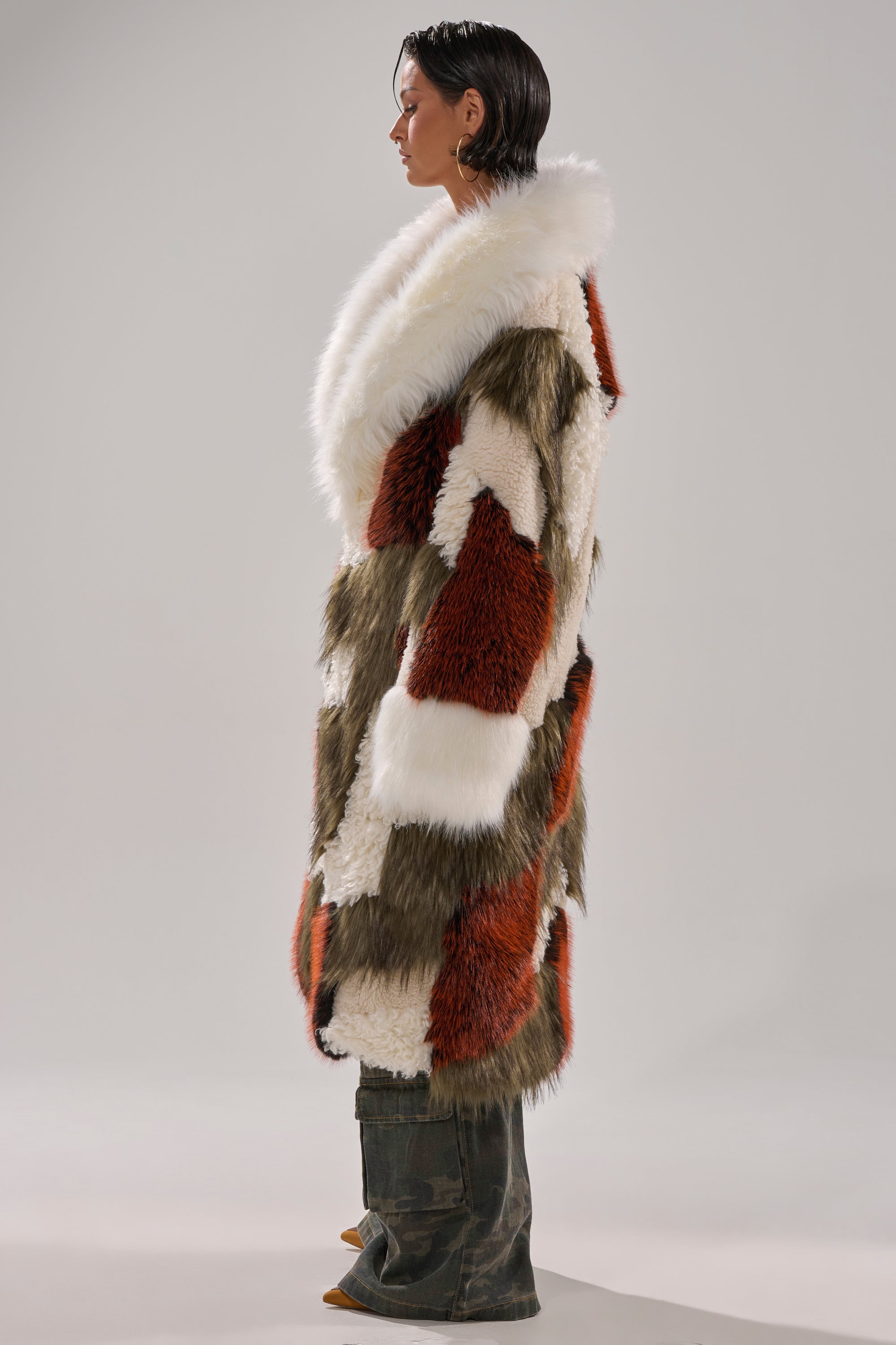 A person stands in profile wearing the FLEUR PATCHWORK FAUX FUR coat with white, brown, and rust fur blocks, paired with wide-legged camouflage pants and pointed shoes against a plain neutral background.