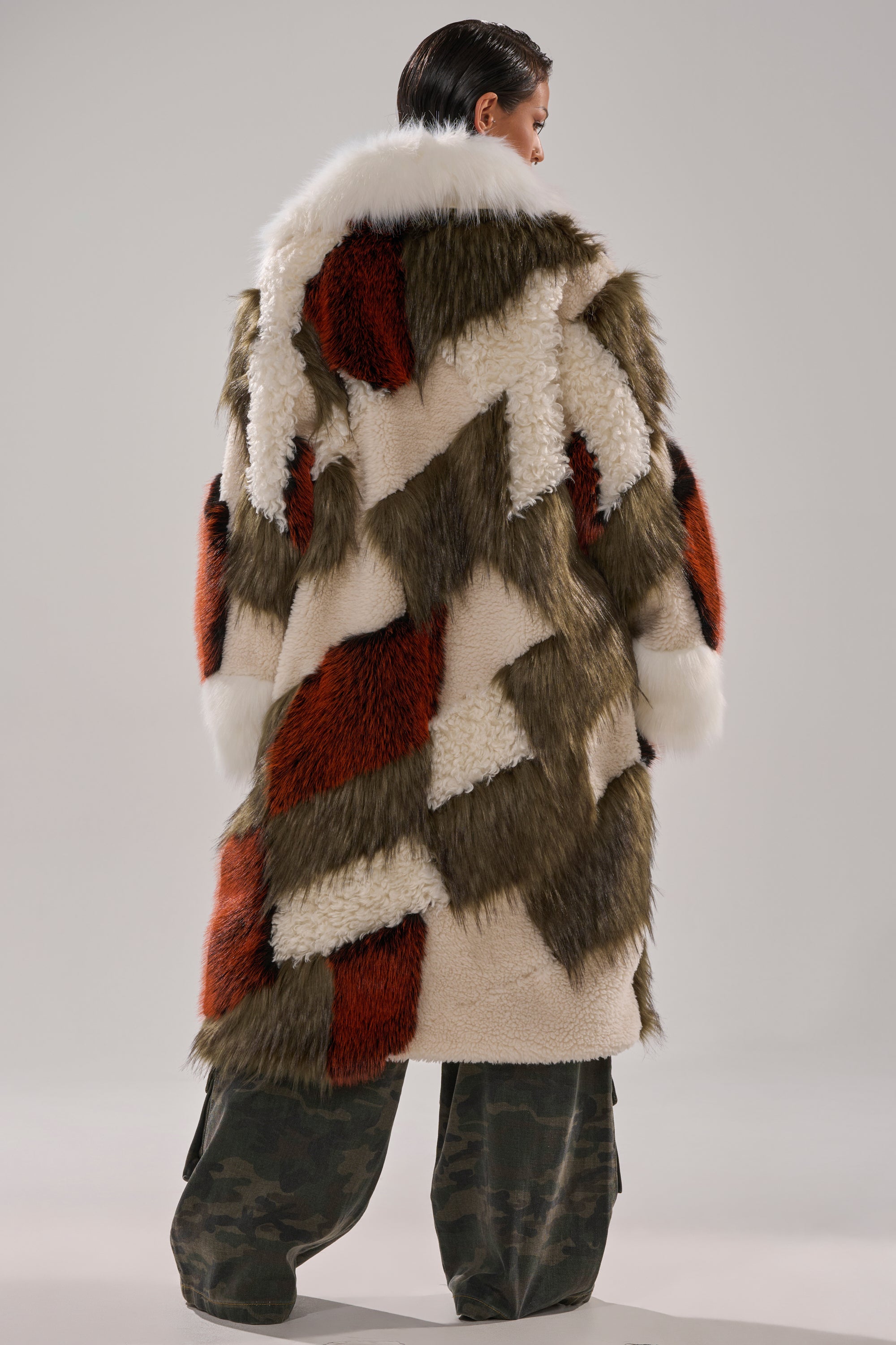 A person wearing the FLEUR PATCHWORK FAUX FUR coat in cream, olive green, and rust tones with wide camouflage pants stands with their back to the camera against a plain background.