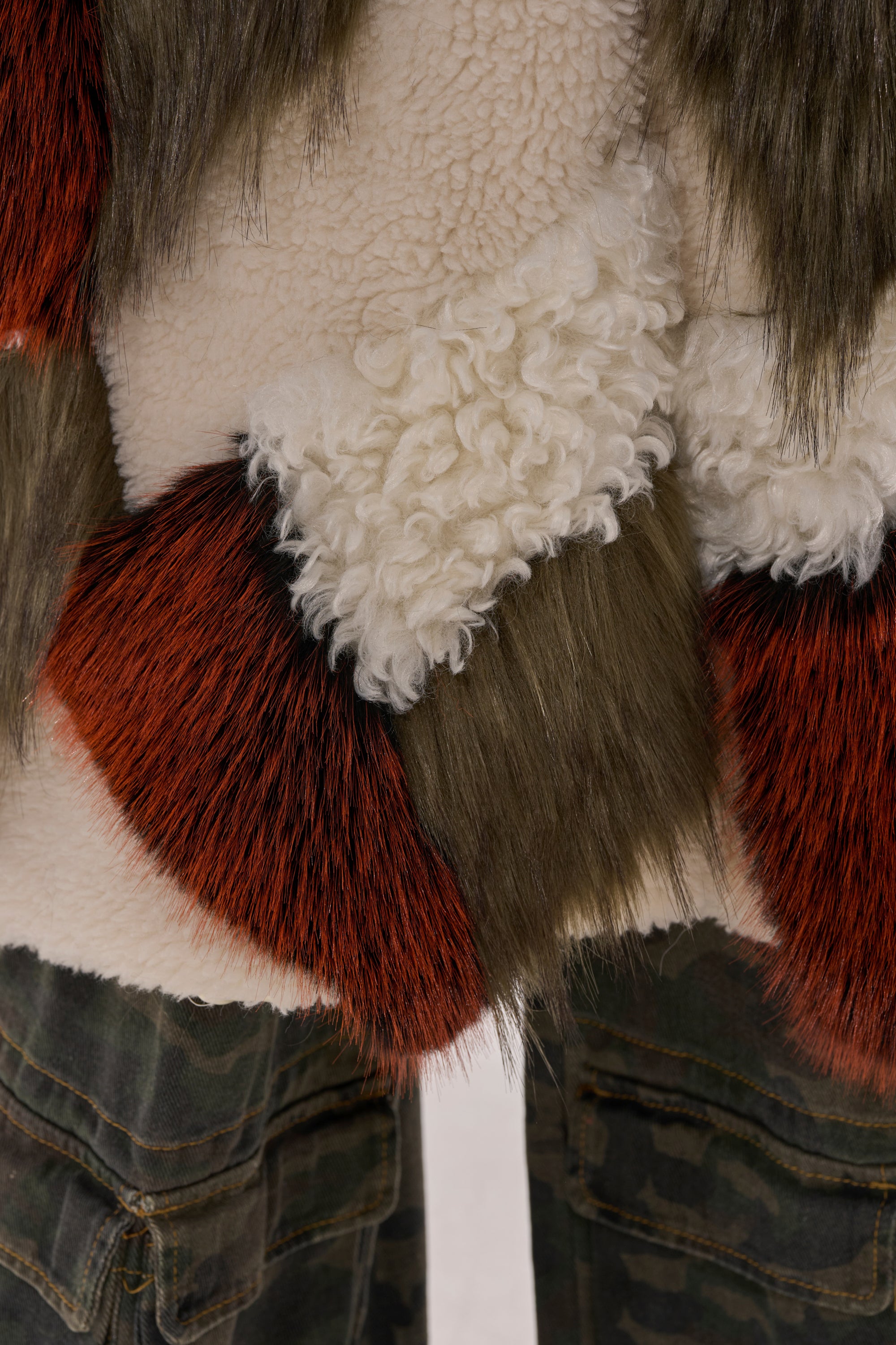 Close-up of FLEUR PATCHWORK FAUX FUR—textured cream shearling with brown and orange fur patches—paired with green camouflage pants at the edge of the image.