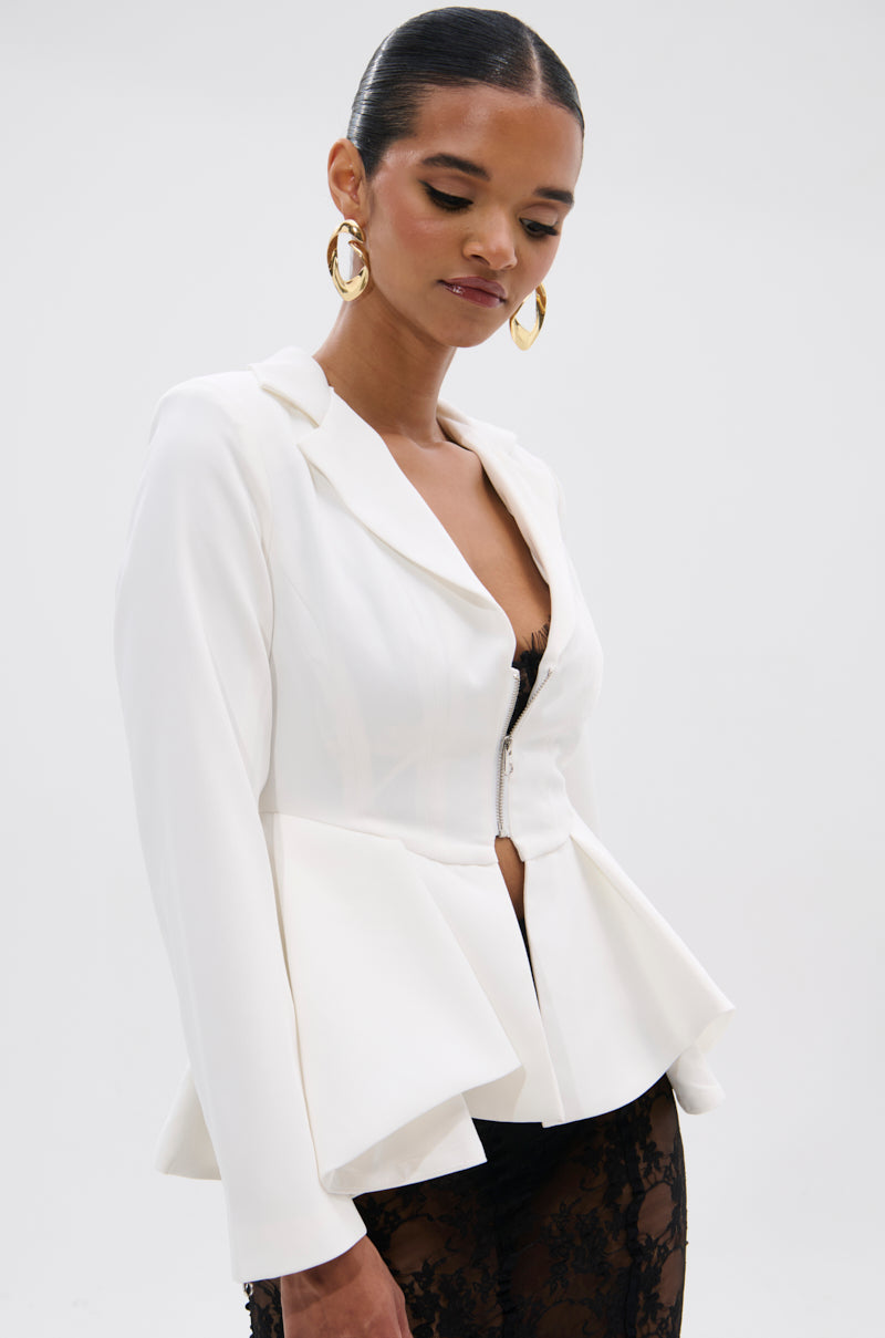 A woman stands against a plain light background, wearing the PARTY IN THE BACK FISHTAIL BLAZER with her hair pulled back and gold hoop earrings, smiling slightly as she looks down.