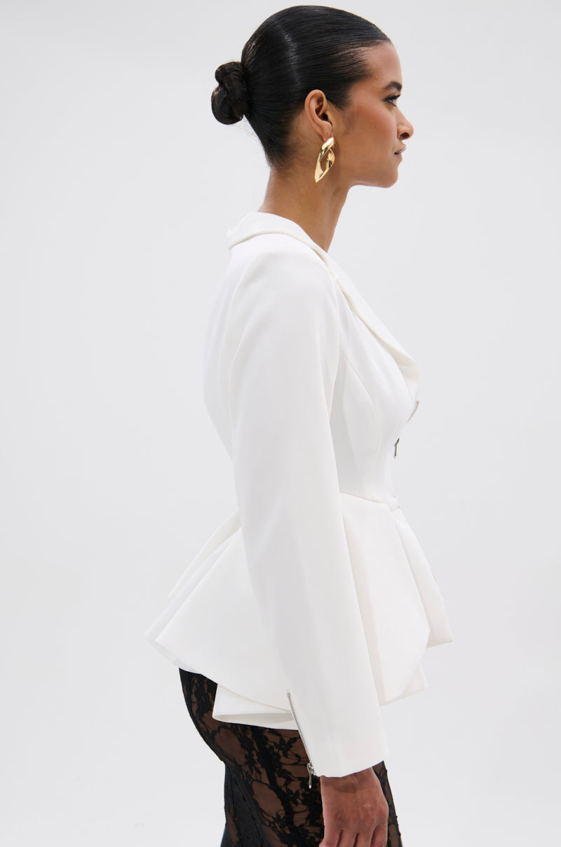 A woman with dark hair in a sleek bun wears the PARTY IN THE BACK FISHTAIL BLAZER in white, styled with black lace pants and large gold earrings, posing in profile against a plain white background.