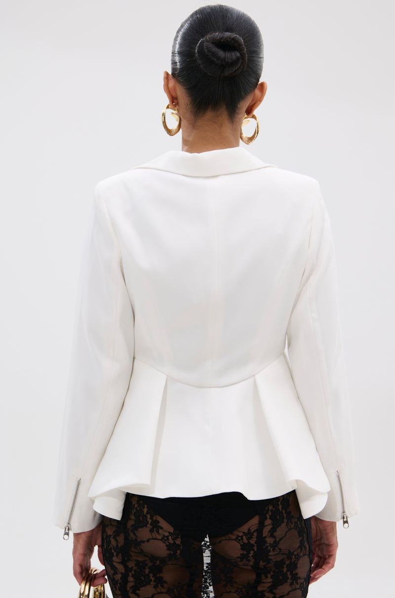 A woman seen from behind wears the PARTY IN THE BACK FISHTAIL BLAZER—a white tailored piece with a peplum hem and zippered sleeves—paired with gold hoop earrings, a sleek bun, and black lace pants.