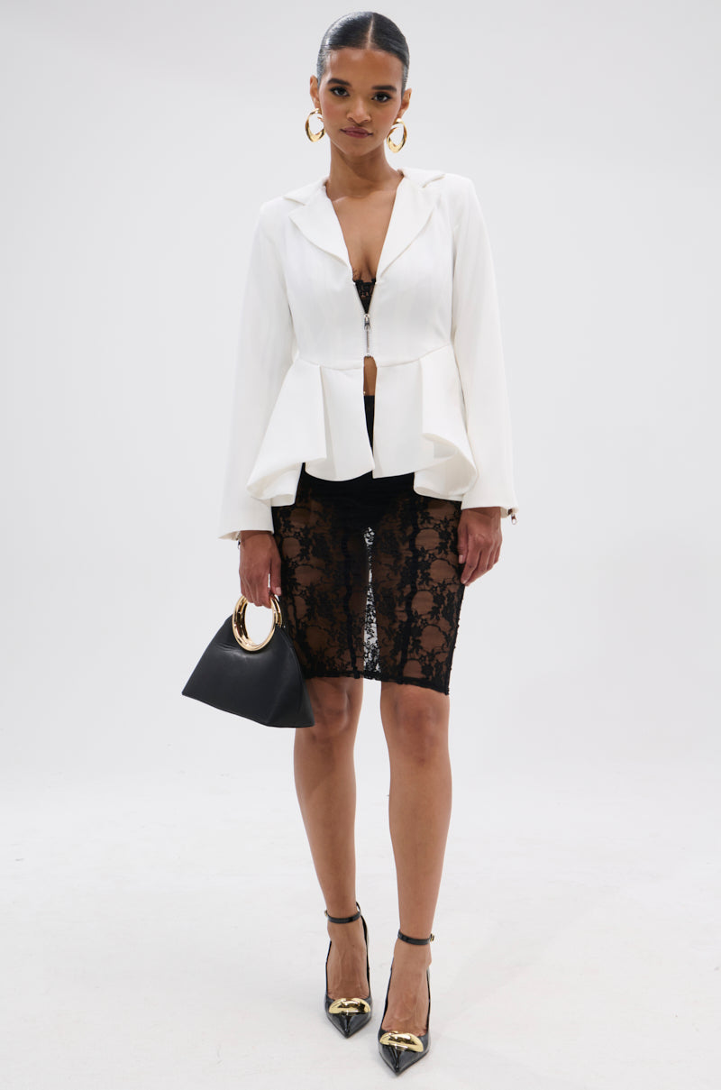 A woman poses against a white background in the PARTY IN THE BACK FISHTAIL BLAZER with a pleated peplum hem, styled over a black lace skirt, gold-toe black heels, and a black handbag with gold handle. She wears hoop earrings and has her hair pulled back.