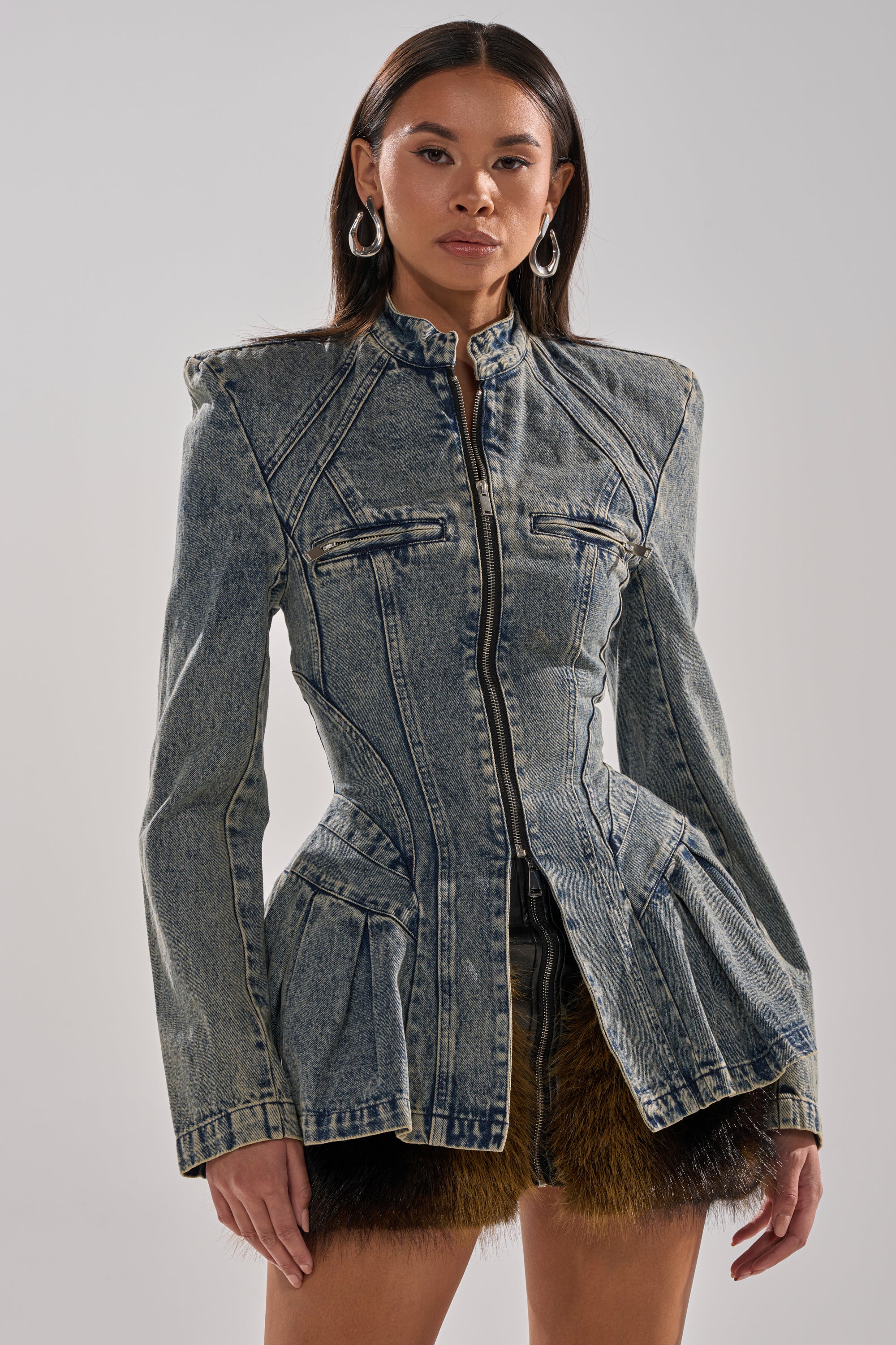 A woman wears the MALLORCA PEPLUM DENIM JACKET with fitted long sleeves, structured shoulders, and a flared hem, styled with a short skirt with dark fur trim, posing against a plain light background.